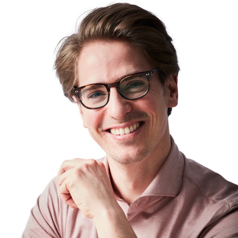 Smiling young man wearing glasses and a light pink shirt, posing with one hand under his chin against a white background.