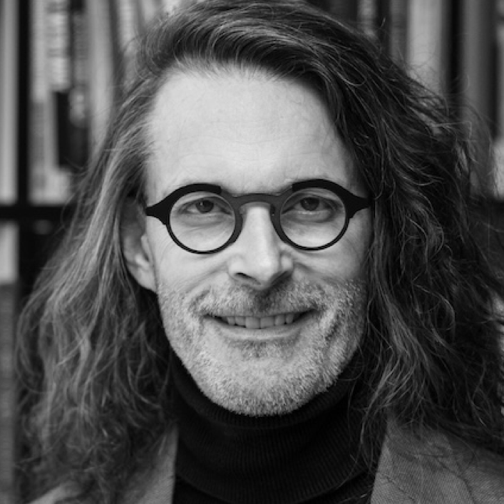 Portrait of a man with long, wavy hair and glasses, smiling confidently in front of a bookshelf. The image is in black and white, highlighting his thoughtful expression and professional demeanor.