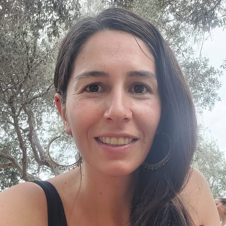 Smiling woman with long dark hair and large hoop earrings, sitting outdoors among trees, enjoying a sunny day.