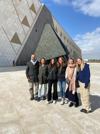 Group of six people posing outdoors in front of a modern architectural building with a geometric design, under a clear blue sky.
