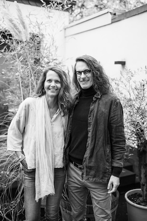 Portrait of a smiling woman and man standing together outdoors, surrounded by greenery, both dressed in casual attire. The image is in black and white, highlighting their friendly expressions and relaxed atmosphere.