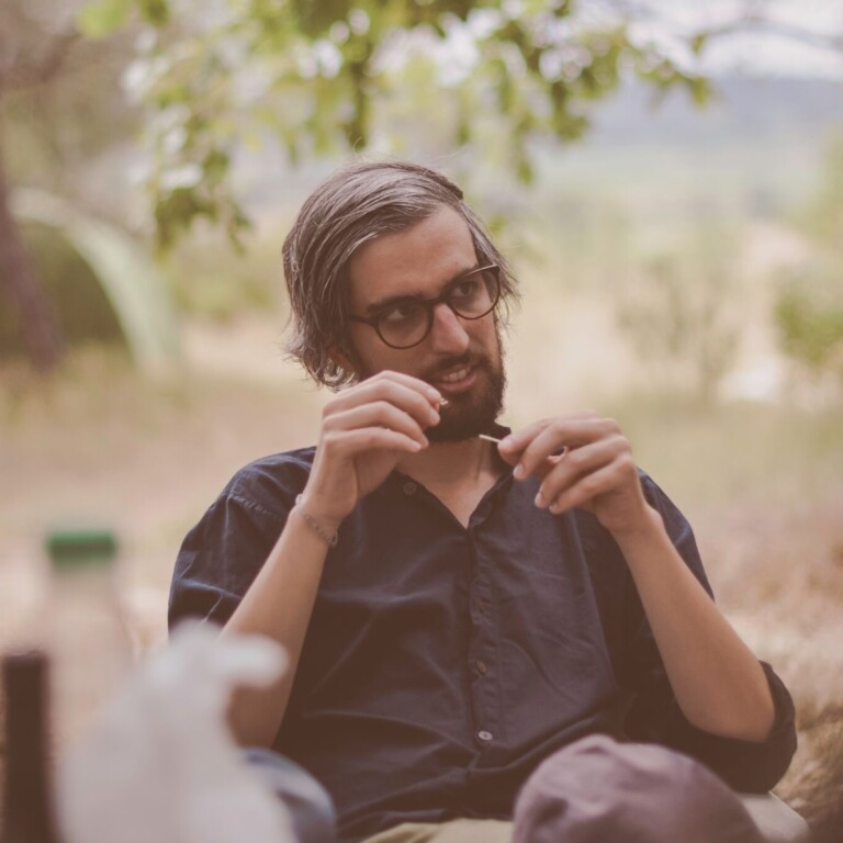 Man with glasses sitting outdoors, holding sunglasses, surrounded by nature.