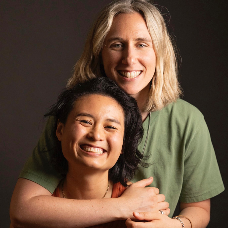 Two smiling women pose together in a warm embrace, showcasing friendship and joy against a dark background. One woman has shoulder-length dark hair and is wearing an orange top, while the other has blonde hair and is dressed in a green shirt. The image captures a moment of happiness and connection.