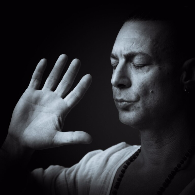 Meditative man with eyes closed, hand raised in a gesture of mindfulness, wearing a simple white garment and beaded necklace, captured in a dramatic black and white setting.