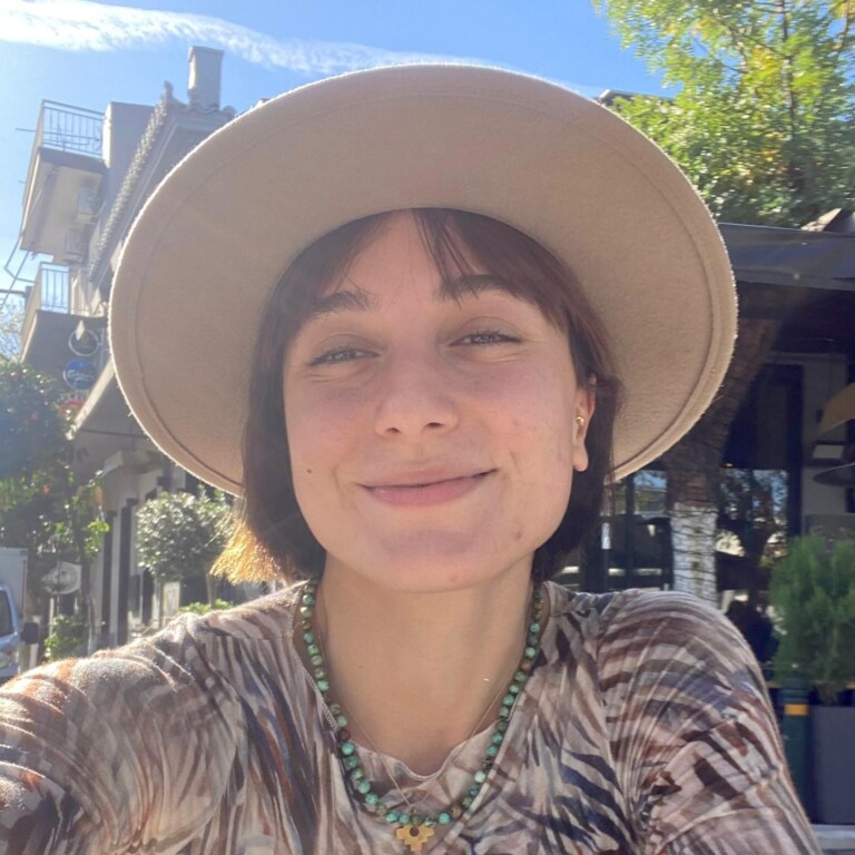 Young woman smiling outdoors, wearing a wide-brimmed hat and a patterned top, with greenery and a sunny sky in the background.